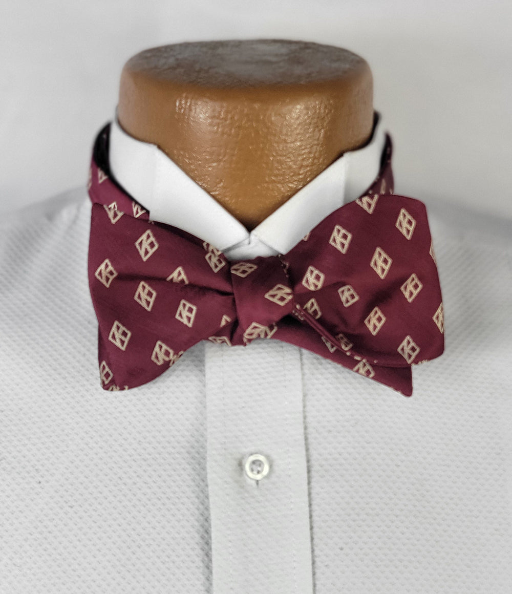 A maroon bow tie with a cream diamond pattern, tied neatly on a white textured dress shirt with a pointed collar, displayed on a light brown mannequin against a neutral light grey background.
Write a product description for the bowtie
Suggest social media posts for the bowtie