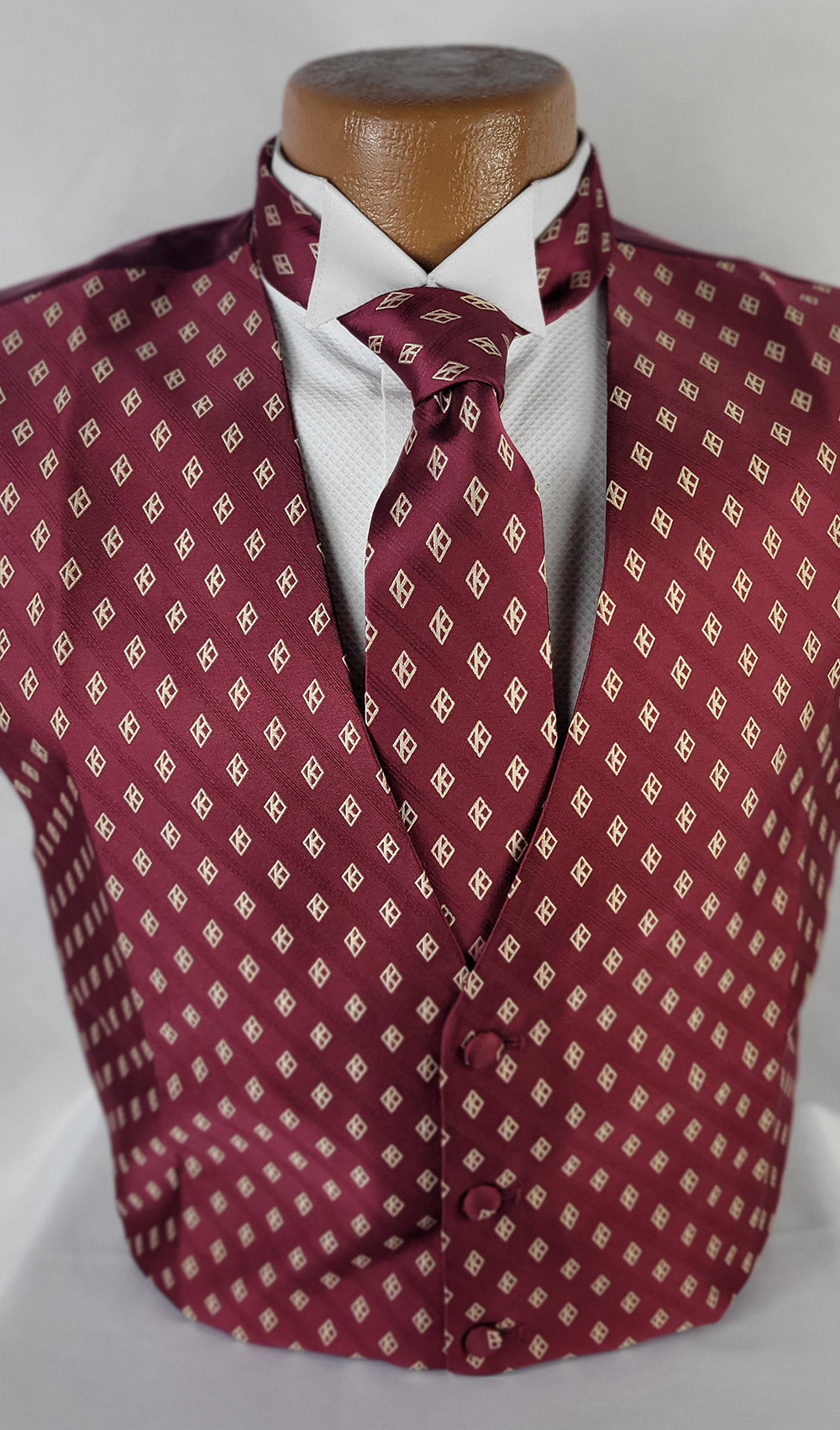 A formal men's wear ensemble featuring a burgundy vest and matching tie with a repeating diamond pattern, displayed on a mannequin over a crisp white dress shirt. The vest has a V-neck opening and three buttons, while the tie is neatly knotted. The background is a plain light color, highlighting the rich burgundy tones of the garments.