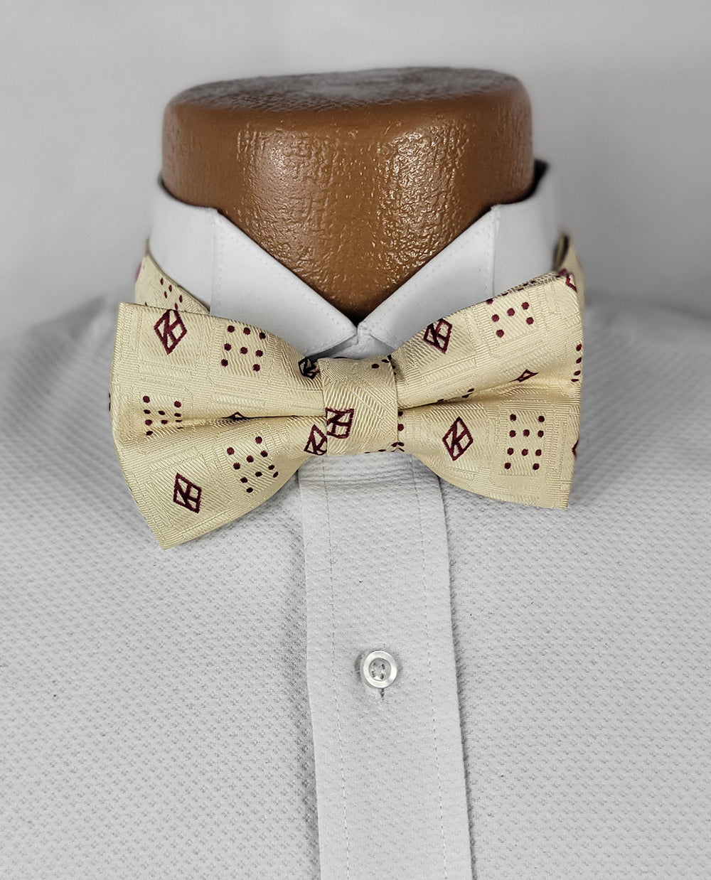 A close-up of the Kappa Founders Floating K Cream Pre-tie Bow Tie, featuring a light cream base with a textured fabric and a unique burgundy pattern of alternating diamond shapes and dot clusters. The bow tie is neatly tied and displayed on a white dress shirt with a crisp collar, set against a neutral gray background.