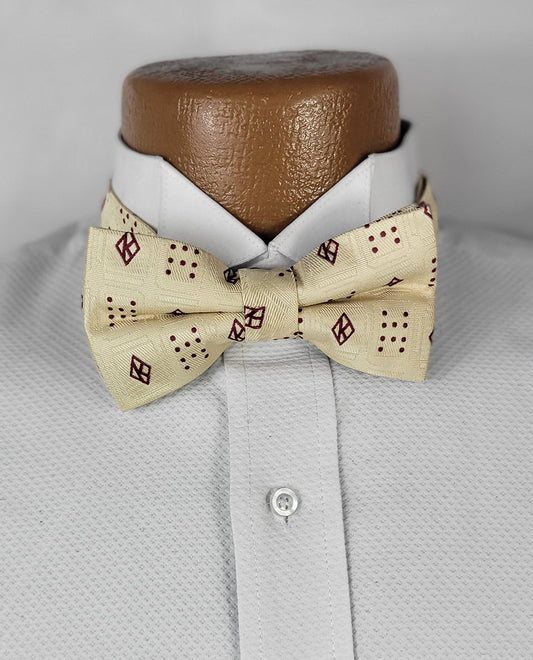 A close-up of the Kappa Founders Floating K Cream Pre-tie Bow Tie, featuring a light cream base with a textured fabric and a unique burgundy pattern of alternating diamond shapes and dot clusters. The bow tie is neatly tied and displayed on a white dress shirt with a crisp collar, set against a neutral gray background.