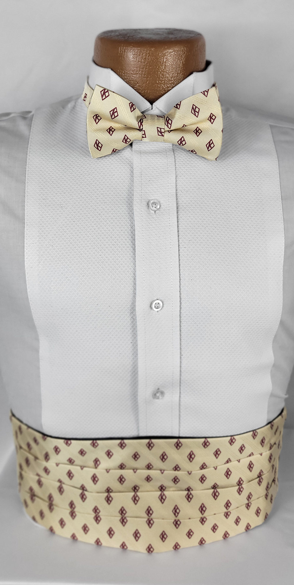 Close-up of a formal menswear ensemble featuring a white tuxedo shirt with a textured front panel, a cream bow tie with burgundy diamond patterns, and a matching pleated cummerbund. Displayed on a dark brown mannequin against a plain white background.