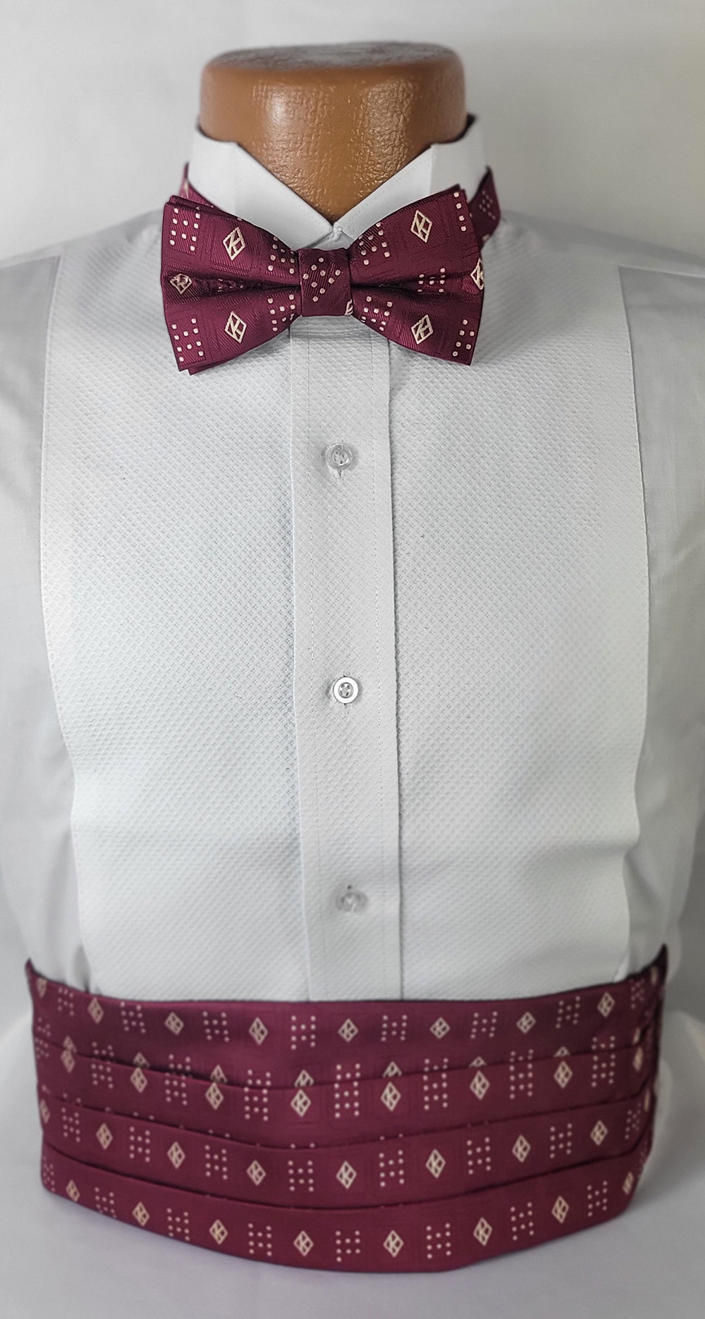 A close-up of a white tuxedo shirt on a mannequin, accessorized with an elegant crimson cummerbund and matching pre-tied bowtie. Both the bow tie and cummerbund feature a sophisticated, repeating geometric pattern.