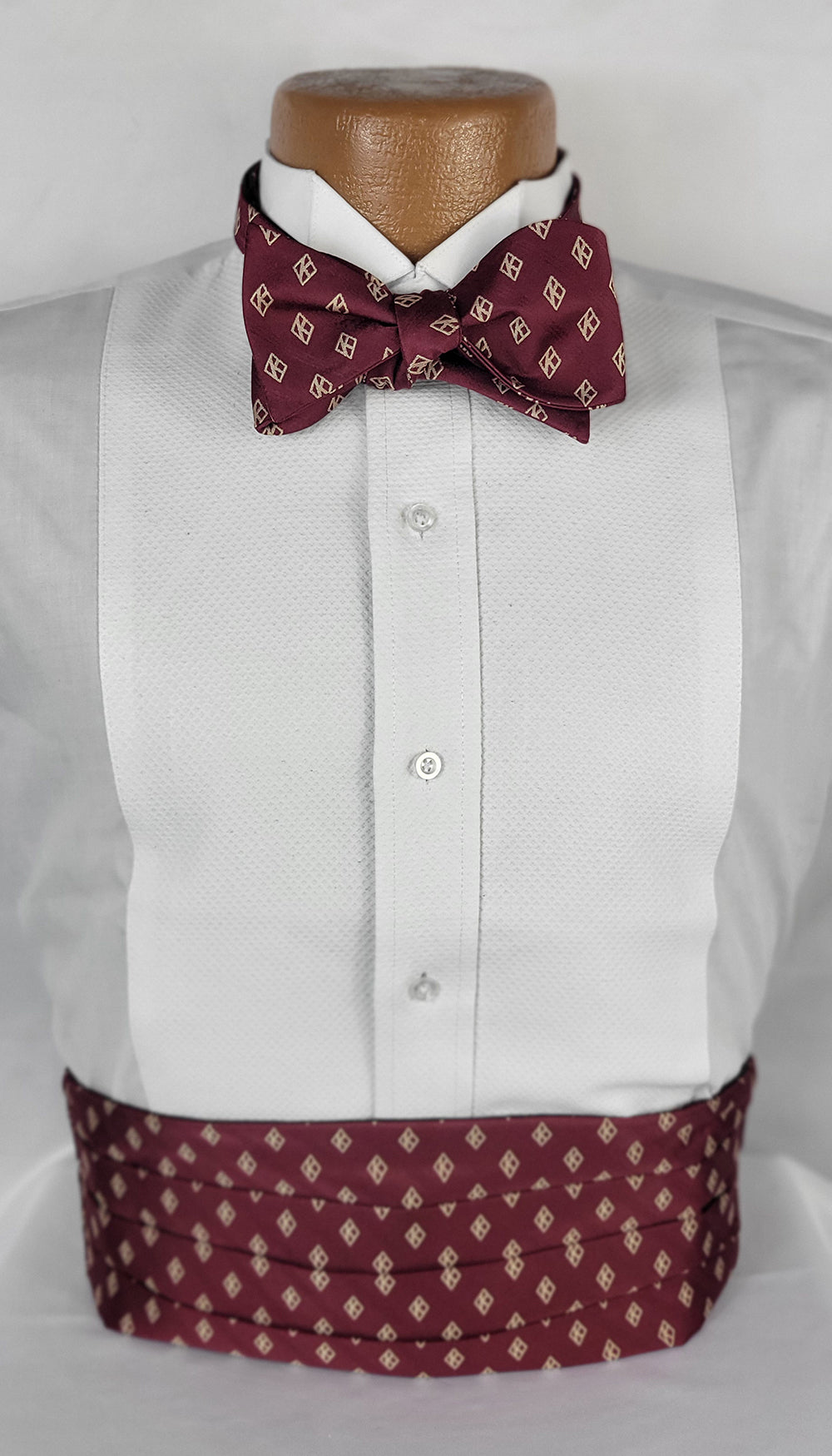 A formal menswear set displayed on a mannequin, featuring a white tuxedo shirt with a textured front panel, a burgundy bow tie with cream geometric patterns, and a matching burgundy cummerbund with pleats and the same pattern. The ensemble is presented against a plain light background.