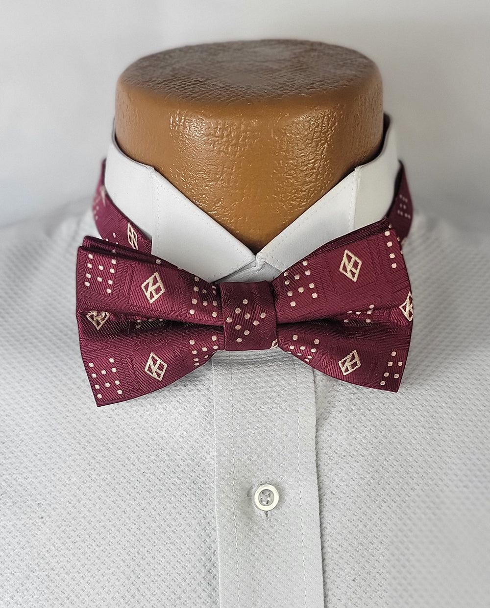A close-up of the Kappa Floating K Founders Crimson Pre-tie Bow Tie, featuring a deep maroon color with a detailed pattern of floating "K" designs, cross-like dot clusters, and diamond-shaped accents. The bow tie is displayed on a white dress shirt with a textured fabric, worn on a brown mannequin bust against a light grey background.