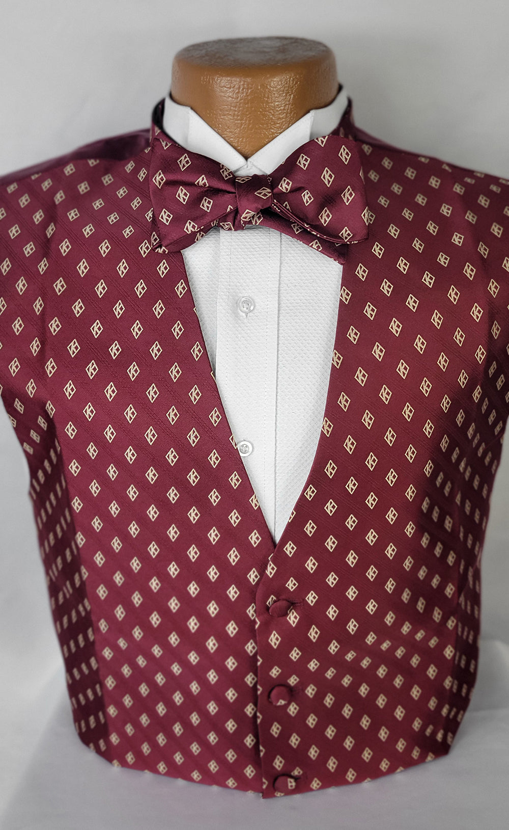 A stylish burgundy vest and matching bow tie with a gold diamond pattern featuring a stylized 'K,' displayed on a mannequin over a white textured dress shirt. Perfect for formal occasions.