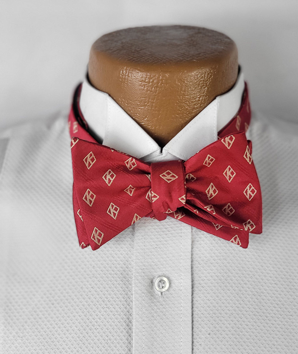 A close-up of a red bow tie with a subtle sheen and a light-colored diamond pattern, neatly tied on a crisp white dress shirt displayed on a brown mannequin. The bow tie exudes sophistication and elegance, perfect for formal occasions.