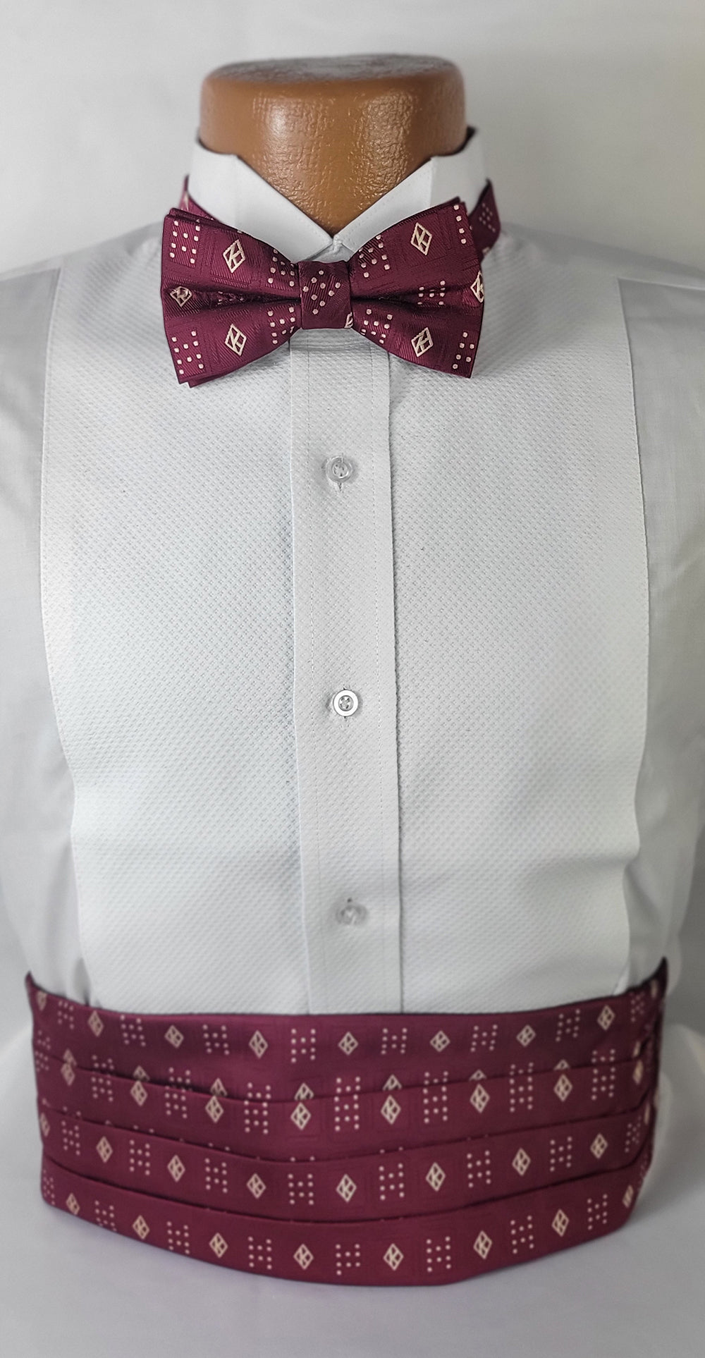 A formal wear display featuring a white tuxedo shirt with a maroon bow tie and matching cummerbund. The bow tie and cummerbund showcase a refined pattern of small white dots and diamond outlines, creating a cohesive and elegant look. The ensemble is presented on a brown mannequin against a plain background.