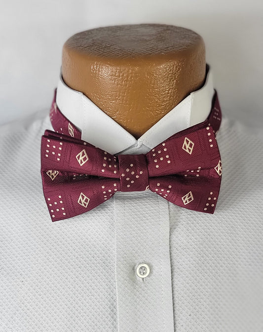A maroon pre-tied bow tie with a distinctive floating 'K' pattern and small dot clusters, displayed on a white dress shirt with a crisp collar, set against a neutral light grey background.