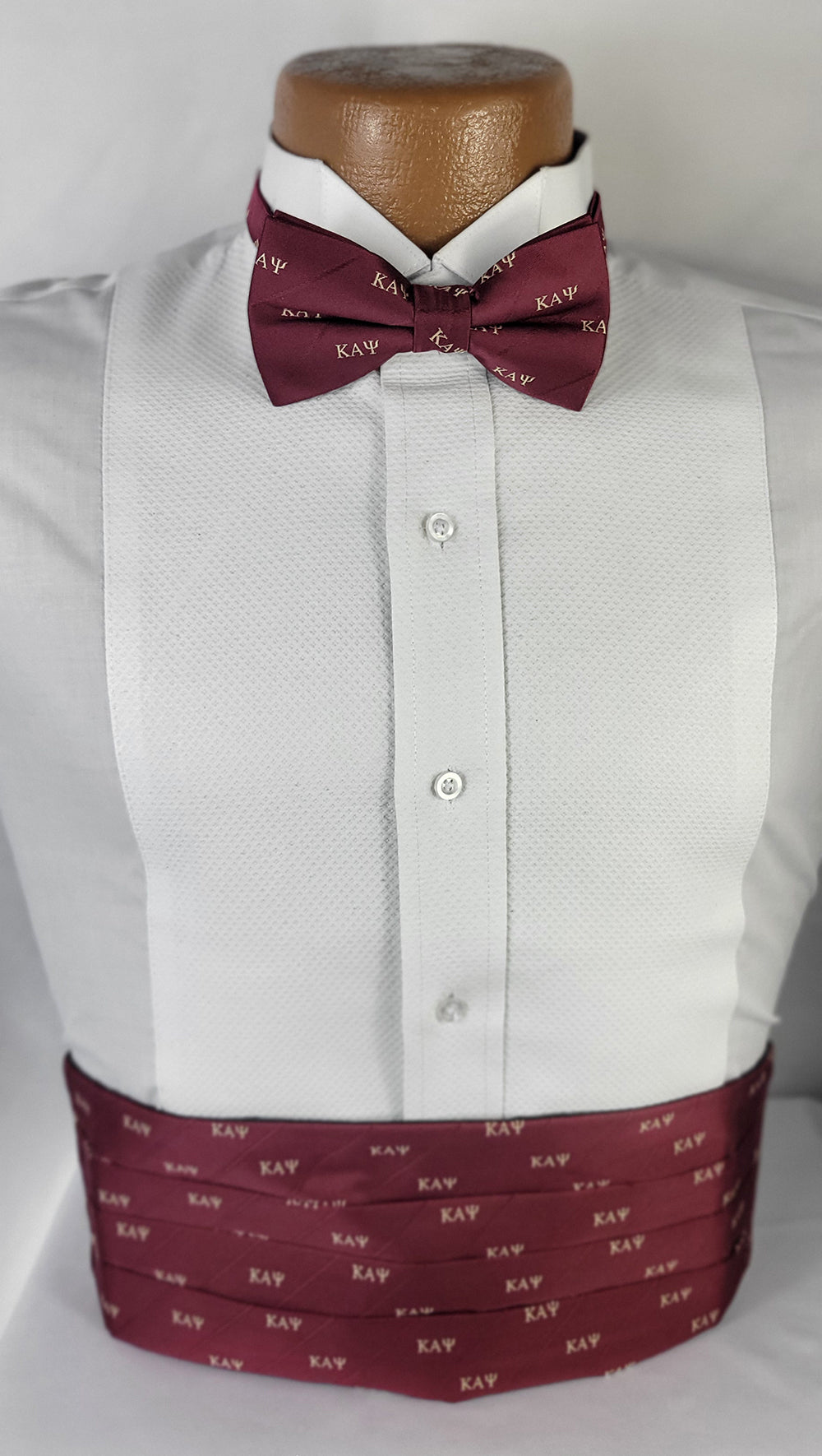 A formal wear ensemble featuring a white tuxedo shirt, a maroon bow tie, and a matching maroon cummerbund, displayed on a mannequin. Both the bow tie and cummerbund are adorned with repeating Greek letters "ΚΑΨ" in a light color, symbolizing Kappa Alpha Psi fraternity. The outfit is set against a plain, light background, emphasizing the elegant and custom design.