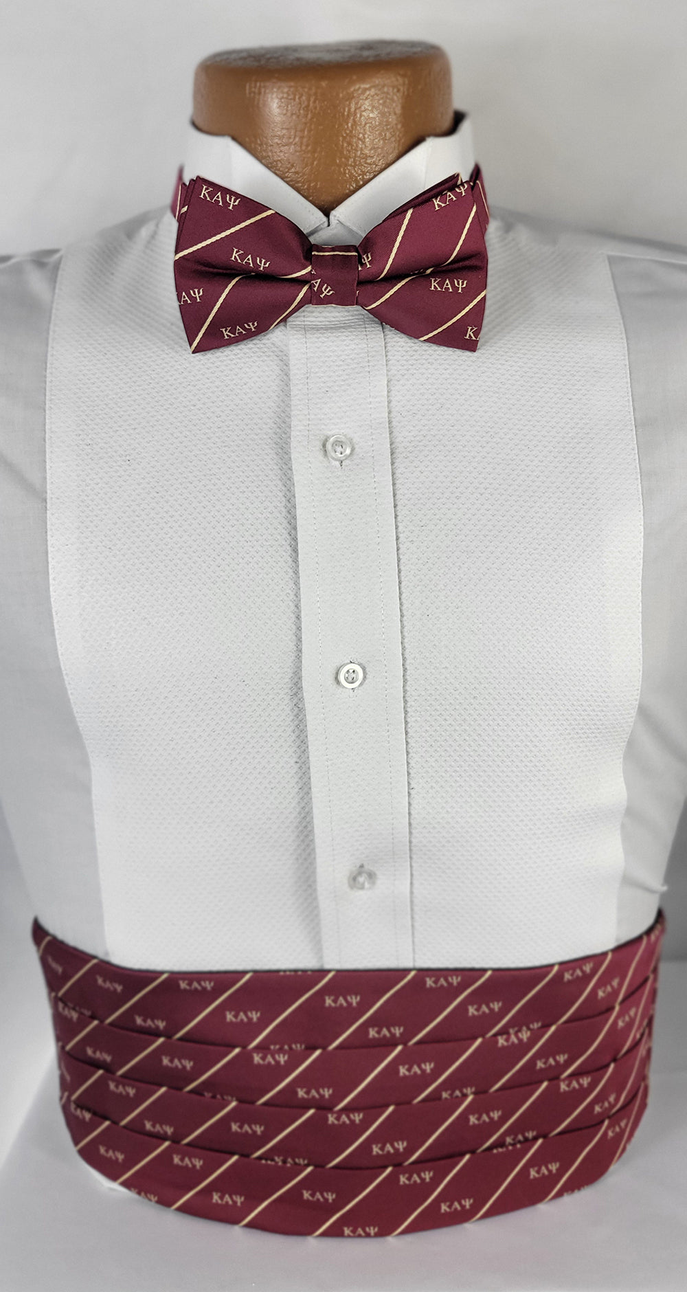 Close-up of a formal Kappa Alpha Psi cummerbund and bow tie set. The maroon accessories feature diagonal cream stripes and repeating 'ΚΑΨ' Greek letters, displayed on a white tuxedo shirt and brown mannequin bust against a light background.