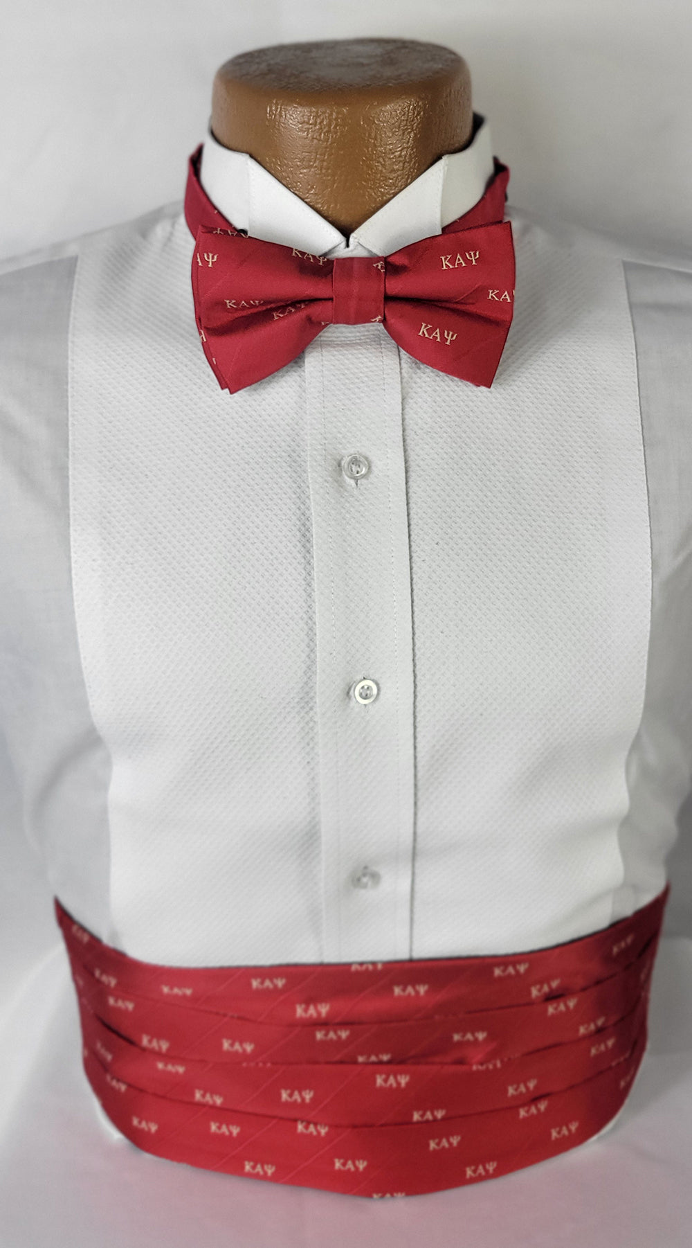 A formal tuxedo shirt displayed on a mannequin, accessorized with a red bow tie and cummerbund. Both accessories feature the Greek letters "KAΨ" (Kappa Alpha Psi) in a light gold color, repeated across the red fabric. The shirt has a textured white pattern, and the ensemble is set against a plain light background.