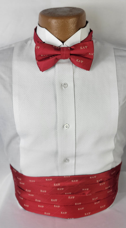 A formal tuxedo shirt displayed on a mannequin, accessorized with a red bow tie and cummerbund. Both accessories feature the Greek letters "KAΨ" (Kappa Alpha Psi) in a light gold color, repeated across the red fabric. The shirt has a textured white pattern, and the ensemble is set against a plain light background.