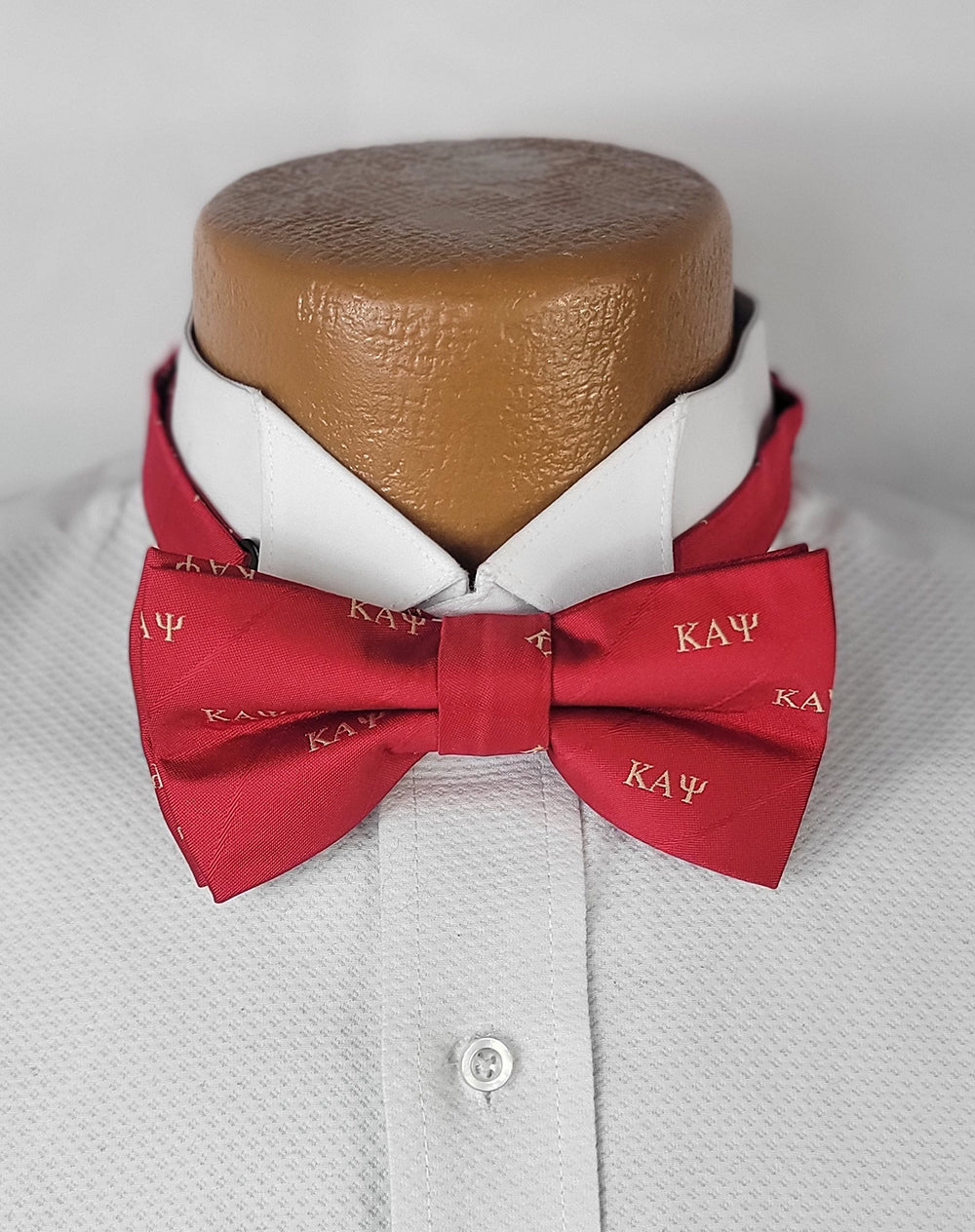 A vibrant red pre-tied bow tie featuring the Greek letters "ΚΑΨ" (Kappa Alpha Psi) in a repeating gold pattern, displayed on a white dress shirt worn by a mannequin torso.